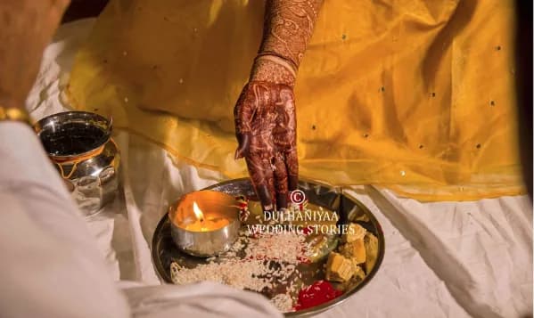 Bride's hennaed hand lighting a diya on a brass haldi thali